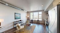 Located at 45 E Reed St in San Jose, CA, this single-floor urban unit presents a contemporary open-concept layout that seamlessly integrates the living, sleeping, and kitchen areas into one cohesive space. A defining architectural detail is the exposed metallic ductwork that runs along the ceiling, lending an industrial-modern character throughout. Wood-look plank flooring extends across the entire unit, providing warmth and visual continuity.

The living area is bright and well-proportioned, anchored by a large picture window that frames views of the surrounding cityscape and invites generous natural light. A built-in window-height counter with bar stools offers an additional casual seating or workspace option. The sleeping area flows naturally from the living space, maintaining the open feel while remaining functional.

The kitchen features light wood cabinetry, a white subway tile backsplash, stainless steel appliances including a refrigerator, range, dishwasher, and microwave, along with open floating shelves for added storage. Countertops are clean and minimal in design.

The bathroom is finished with a light wood vanity, an undermount sink with white countertop, a large wall mirror, and a walk-in shower, all set atop wood-look flooring that matches the rest of the unit. The overall ambiance is clean, modern, and urban.