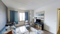 This single-floor space showcases a thoughtfully designed living room that balances comfort and style. The room features hardwood-style flooring throughout, anchored by a patterned area rug that defines the seating area. A large window dressed with teal grommet curtains allows natural light to fill the space, creating a bright and welcoming atmosphere.

The living area is furnished with a coordinated seating arrangement, including a sofa and accent chair, centered around a sleek white coffee table. A media console along the far wall houses a flat-screen television and is complemented by woven storage baskets, offering both functionality and visual interest. Above the media setup, a large multi-panel abstract artwork in soft blues and neutrals serves as a prominent decorative focal point.

The walls are finished in a neutral tone, and the textured ceiling adds subtle architectural character. A floor lamp with a tripod base provides warm supplemental lighting. The overall ambiance is casual yet curated, with a cohesive color palette of blues, grays, and warm wood tones that lends the space a relaxed, contemporary feel.
