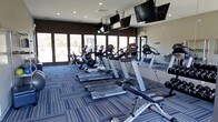 This fitness center offers a thoughtfully designed workout environment that combines functionality with a clean, contemporary aesthetic. The space is anchored by a lineup of Cybex cardio equipment, including treadmills, ellipticals, and a recumbent bike, arranged to maximize both usability and flow. Large windows along one wall flood the room with natural light and provide open views of the surrounding outdoor landscape, creating an energizing atmosphere for exercise. The flooring features a modern patterned carpet in cool blue and grey tones, adding a polished, professional look underfoot. Full-length mirrors run along one side of the space, visually expanding the room and allowing users to monitor their form during workouts. Multiple flat-screen TVs are suspended from the ceiling and mounted on the walls, ensuring entertainment and information are accessible from virtually any point in the room. Recessed lighting in the ceiling complements the natural light, keeping the space bright and well-illuminated at all times. The overall ambiance is clean, focused, and inviting, making it a well-rounded facility suited for a variety of cardio and fitness routines.