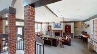 This single-floor community clubhouse is thoughtfully designed to serve as a comfortable and inviting gathering space. The interior showcases a blend of classic and contemporary design elements, anchored by prominent brick columns and exposed beam accents that add architectural character throughout.

The main lounge area is the heart of the space, featuring soaring vaulted ceilings that lend an open, airy feel. A handsome brick fireplace serves as the room's focal point, flanked by built-in shelving units with warm accent lighting. Plush leather seating, a patterned area rug, and a flat-screen television mounted above the fireplace create a relaxed, home-like atmosphere.

A split-level layout adds visual interest, with steps and black metal railings defining distinct zones within the open floor plan. One area is arranged as a casual seating nook with accent chairs, while another features counter-height bar seating positioned beside oversized grid-pane windows that flood the space with natural light and provide views of the surrounding pool area.

French doors open directly to the outdoor pool area, further connecting the interior to the exterior amenity spaces. Soft patterned carpeting, recessed lighting, and a warm neutral color palette with red accents complete the welcoming ambiance of this well-appointed clubhouse.