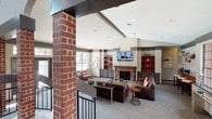 This single-floor community clubhouse is thoughtfully designed to serve as a comfortable and inviting gathering space. The interior showcases a blend of classic and contemporary design elements, anchored by prominent brick columns and exposed beam accents that add architectural character throughout.

The main lounge area is the heart of the space, featuring soaring vaulted ceilings that lend an open, airy feel. A handsome brick fireplace serves as the room's focal point, flanked by built-in shelving units with warm accent lighting. Plush leather seating, a patterned area rug, and a flat-screen television mounted above the fireplace create a relaxed, home-like atmosphere.

A split-level layout adds visual interest, with steps and black metal railings defining distinct zones within the open floor plan. One area is arranged as a casual seating nook with accent chairs, while another features counter-height bar seating positioned beside oversized grid-pane windows that flood the space with natural light and provide views of the surrounding pool area.

French doors open directly to the outdoor pool area, further connecting the interior to the exterior amenity spaces. Soft patterned carpeting, recessed lighting, and a warm neutral color palette with red accents complete the welcoming ambiance of this well-appointed clubhouse.