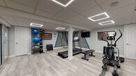 This well-appointed single-floor space seamlessly blends a professional fitness facility with functional office areas. The open gym zone is generously sized, featuring light wood-look flooring throughout that lends a clean, contemporary aesthetic. A suspended drop ceiling houses striking geometric LED light panels that provide bright, even illumination across the entire space.

The fitness area is comprehensively equipped, including a treadmill, elliptical trainer, cable machine, weight bench, dumbbell rack, and a standing ball rack stocked with medicine balls and stability balls of various sizes. A wall-mounted flat-screen television serves the workout zone, while the accent wall features bold teal and grey geometric designs that add visual energy to the space.

Transitioning toward the office zone, the space offers multiple workstations with desktop computers and ergonomic seating. A set of glass-partitioned private rooms with frosted lower panels provides semi-private areas suitable for consultations or focused work, maintaining an open feel while offering separation. A doorway leads to an exterior staircase, providing direct access to the outside. White structural columns are distributed throughout, complementing the neutral grey walls and cohesive modern finishes.