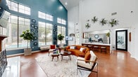 Located at 2323 Eldridge Parkway in Houston, TX, this thoughtfully designed single-floor space makes a striking first impression with its soaring double-height ceilings and bold architectural character. The living room is the centerpiece of the layout, showcasing dramatic teal accent walls that rise to meet a bank of oversized clerestory windows, flooding the space with abundant natural light. Views of a pool area can be seen through the floor-to-ceiling windows, adding an inviting outdoor connection. A classic wood-paneled fireplace with built-in cabinetry anchors one wall, providing a warm focal point complemented by warm terracotta-toned polished concrete flooring that runs throughout. Curated wall art and lush indoor plants enhance the mid-century modern aesthetic. The kitchen is efficiently arranged in an L-shaped layout, featuring white cabinetry, light-toned countertops, and stainless steel appliances including a full-size refrigerator and dishwasher. A window above the sink brings in natural light, while hanging planters add a fresh, organic touch. The overall ambiance of the space is vibrant yet welcoming, blending bold design choices with functional, open living areas ideal for both gathering and everyday use.