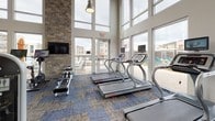 Located at 6600 Rolling Meadows Dr in Sparks, Nevada, this fitness center is a thoughtfully designed, single-floor facility built to support a comprehensive workout experience. The main exercise floor is defined by soaring ceilings with a wood-paneled sloped finish and exposed ductwork, giving the space an open, industrial-modern character. Floor-to-ceiling windows line multiple walls, flooding the interior with natural light and offering views of the surrounding outdoor pool area and community grounds.

The space is well-appointed with a wide array of strength training machines, a dedicated free weights section featuring a full dumbbell rack with mirrored backing, a power rack, and multiple adjustable benches. The cardio zone includes treadmills and rowing machines positioned along the window walls to take advantage of the outdoor views, alongside elliptical trainers and additional cardio equipment. A mounted flat-screen TV provides entertainment throughout.

A separate functional training annex connects to the main floor through a large garage-style roll-up door, creating flexible indoor-outdoor access. This area is equipped with a punching bag, battle ropes, medicine balls, and resistance training accessories, complemented by turf access for dynamic movement exercises. Patterned carpet tiles in blue and tan tones ground the space throughout, contributing to the modern aesthetic of this well-rounded fitness facility.