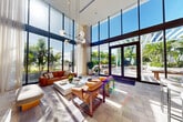 Located at 247 NE 29th St in Miami's vibrant Edgewater district, this elegantly designed lounge space makes a striking architectural statement with its dramatic double-height ceilings and expansive floor-to-ceiling glass walls. Abundant natural light pours in throughout the day, illuminating the refined stone tile flooring and creating a seamless visual connection to the lush tropical landscaping and outdoor terrace beyond.

The interior is thoughtfully curated with a sophisticated yet inviting aesthetic. A bold botanical-print accent wall serves as a dramatic focal point, framing a large flat-screen display and anchoring the main seating area. Rich cognac leather sofas and mid-century-inspired wooden armchairs are arranged around a white stone coffee table, creating an inviting conversation area. A built-in bar counter with sleek bar seating lines one wall, accented by a luminous green glazed tile backsplash that adds a vibrant, artistic touch.

Statement globe pendant lighting descends from the soaring ceiling, adding warmth and a modern sculptural quality to the space. Large sliding glass doors open directly to a sunlit outdoor terrace surrounded by mature palms, extending the living experience to the outdoors. The overall ambiance is light, airy, and distinctly Miami — blending contemporary design with a lush, tropical sensibility.