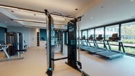 This contemporary fitness center is thoughtfully designed to support a comprehensive range of workout styles across a single, spacious floor. The facility is divided into distinct functional zones, each outfitted with professional-grade equipment. The cardio area lines the perimeter near expansive floor-to-ceiling windows, where rows of treadmills, ellipticals, and stationary bikes are positioned to take full advantage of natural light and views of the surrounding landscaped outdoor areas and urban surroundings. A dedicated strength training zone features multiple Precor resistance machines arranged to allow comfortable movement between stations, complemented by a cable crossover system for versatile strength work. A separate functional training studio offers a more intimate setting, equipped with suspension training rigs, yoga mats, and mirrored walls that amplify the sense of space. Framed artwork along the walls adds a refined aesthetic touch to this area. Throughout the facility, a neutral, light-toned floor provides a clean and durable surface, while recessed and linear pendant lighting creates a bright, energizing atmosphere. Teal accent walls add a distinctive visual character, grounding the modern design. Built-in storage and shelving units offer convenient access to accessories, rounding out this well-appointed fitness destination.