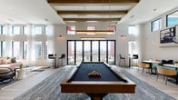 Located at 2600 N Orange Ave in Orlando, this single-floor amenity space is thoughtfully designed to blend social and recreational functions within a modern, open-concept layout. The space is defined by soaring ceilings with exposed wood beams, floor-to-ceiling windows, and an abundance of natural light that frames panoramic views of the Orlando skyline throughout.

The lounge area is anchored by a plush seating arrangement featuring a pink sectional sofa, leather accent chair, and sculptural coffee tables atop a bold striped area rug. A wall-mounted television and built-in shelving provide both function and visual interest. Pendant lighting adds warmth and character overhead.

The games area centers on a full-sized billiards table set beneath dramatic coffered ceilings with exposed beams. High-top bistro tables line the perimeter, and large glass doors open directly onto an outdoor terrace with sweeping city views.

A well-appointed wet bar features green subway tile, open wood shelving, a undermount sink, and stainless steel beverage refrigerators. A dark-toned dining table with woven chairs bridges the lounge and bar areas. An adjacent glass-door room offers a semi-private workspace. Light wood-look flooring flows throughout, unifying the entire space.