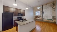 This single-floor unit blends modern finishes with distinctive industrial character. The open-concept kitchen serves as the focal point, featuring a central island with granite countertops and an integrated sink, dark espresso cabinetry, and black appliances including a refrigerator, electric range, and microwave. Pendant lights hang from the elevated ceilings, which also display exposed ductwork and piping, reinforcing the loft-inspired aesthetic.

Warm hardwood flooring extends throughout the main living area, complementing the rich cabinetry tones and neutral gray walls. A standout architectural detail is the exposed brick and weathered plaster accent wall, which adds raw, historic texture to the space and serves as a conversation piece within the living area. A window on this wall allows natural light to filter in, while a ceiling fan provides additional airflow.

A separate room with continued hardwood flooring and its own window offers flexible use as a bedroom or additional living space. The bathroom is cleanly finished with tile flooring, a granite-topped dark wood vanity, a full tub-shower combo, and a closet, providing a polished contrast to the unit's more industrial elements.