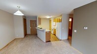 This single-floor residence offers a functional and welcoming layout centered around an open-concept living and kitchen area. The kitchen is outfitted with honey-toned wood cabinetry that extends to the upper walls, providing ample storage space. White appliances, including a refrigerator, electric range, and microwave, complement the clean and practical design. A peninsula with a light-colored countertop extends into the living area, creating a natural divide between the kitchen and the carpeted main living space while also offering additional prep or casual dining surface.

The flooring transitions from warm hardwood-style planks in the kitchen to plush, neutral-toned carpet in the living area, adding comfort underfoot. The walls are painted in a soft, warm neutral tone that contributes to the inviting atmosphere throughout. A solid wood entry door with a classic six-panel design provides a sturdy and traditional entryway. Doorways visible from the main space lead to additional rooms, including what appears to be a bathroom, offering hints of the full layout beyond the central living area. Overall, the space presents a practical, comfortable, and well-organized residential environment.