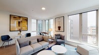 This contemporary single-floor residence offers a thoughtfully designed layout that balances comfort and modern style. The living area is a standout feature, bathed in natural light streaming through large corner windows that frame urban views. The space is appointed with stylish furnishings, including a plush sectional sofa, coordinating accent chairs, and round white coffee tables, all set atop a blue area rug. Framed artwork and warm lighting add character to the clean white walls.

The kitchen is outfitted with dark wood-grain cabinetry, stainless steel appliances — including a range, microwave, and dishwasher — light stone countertops, and a classic white subway tile backsplash. Light hardwood flooring runs throughout, lending warmth and continuity across the space.

The bedroom is serene and minimalist, featuring crisp white bedding with patterned accent pillows, a wall-mounted climate control unit, and a large sliding glass door that allows natural light to filter in from the surrounding urban environment.

The bathroom is finished with dramatic dark rectangular wall tiles surrounding a clean white soaking tub, creating a sleek, contemporary aesthetic. Overall, the residence presents a polished, modern character with a cohesive design palette throughout.