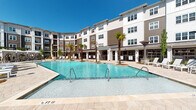 Located at 7770 Market St in Wilmington, North Carolina, this multi-story residential community is designed around a striking central courtyard that serves as the focal point of the property. The architecture blends neutral tones of white, taupe, and warm brown with horizontal siding and large windows, creating a modern coastal aesthetic that complements the Wilmington climate.

The outdoor amenity area is a standout feature, centered on a generously sized resort-style swimming pool with crystal-clear blue water and a stone-tiled surround. Manicured landscaping lines the pool deck, with young shade trees, ornamental shrubs, and mulched planting beds framing rows of white lounge chairs. The open-air courtyard is designed for both relaxation and social gathering, with covered pergola structures and a covered outdoor dining area overlooking the pool, allowing residents to enjoy the surroundings in shade.

Interior common areas feature tile flooring in a diagonal pattern and clean white walls accented by distinctive dark slatted wood partitions, lending a contemporary, well-organized feel to the corridors. Natural light filters through at corridor ends, connecting interior passages to the outdoor courtyard beyond. The overall ambiance of the community is bright, polished, and resort-inspired.