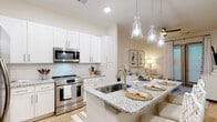 Located at 6146 Liberty Hall Dr in Leland, NC, this single-floor residence offers a thoughtfully designed open-concept layout filled with clean lines and a light, coastal-inspired aesthetic. The kitchen is a standout feature, showcasing crisp white shaker cabinetry, granite countertops, a subway tile backsplash, and a full suite of stainless steel appliances including a range, over-the-range microwave, and French door refrigerator. A granite-topped island with seating for multiple provides both a casual dining space and a natural focal point, illuminated by elegant glass pendant lights. Light wood-style flooring flows throughout the main living areas, enhancing the sense of openness. The living area is warm and inviting, with access to an outdoor balcony through a sliding glass door that invites natural light and outdoor views. The bedroom is a serene retreat featuring plush carpet, a wall-mounted television, and generous natural light from well-dressed windows. The en-suite bathroom is finished with granite countertops, white cabinetry, a glass-enclosed walk-in shower, and wood-style flooring. A spacious walk-in closet with wire shelving and hanging storage provides ample organization. The overall ambiance is bright, clean, and refined throughout.