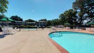 Located at 986 Armfield Circle in Norfolk, Virginia, this single-floor community amenity space offers a well-appointed outdoor recreation area paired with a neatly maintained interior entryway corridor. The outdoor area is the centerpiece of this space, featuring a generously sized swimming pool with gently stepped entry points and brick-accented coping, alongside a separate shallow wading pool — ideal for all ages. The pool deck provides ample open space for lounging, complemented by patio chairs and umbrellas for shade. The area is enclosed by a security fence and is framed by mature trees that offer natural shade and a lush, green backdrop under open skies. The interior corridor connects residents to the amenity areas and is finished with earthy, multi-toned slate-style tile flooring that adds a warm, grounded aesthetic. Cream-colored walls and crisp white millwork keep the space feeling clean and bright. Coastal-themed framed artwork — depicting sailboats on calm water and a vibrant pier at sunset — reinforces the nautical character of the Norfolk waterfront community. Safety features including a fire alarm pull station are clearly visible, reflecting a commitment to resident well-being. Together, the outdoor and interior elements create a cohesive, inviting community environment.