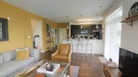 This single-floor residence at Penn Circle Apartments offers an open-concept layout that integrates the living and kitchen areas into one cohesive, functional space. The living area features rich hardwood flooring, warm mustard-yellow accent walls, and a decorative brick column that adds an industrial-chic touch. A lean ladder shelf provides both style and storage, while large windows with white blinds allow natural light to fill the room. The seating arrangement, anchored by a leather accent chair and a glass coffee table, creates a comfortable and well-appointed gathering space. The kitchen is defined by dark cabinetry that contrasts sharply with a light granite or stone-topped breakfast bar, which is lined with modern white bar stools. Three pendant lights hang above the bar, providing focused task lighting and visual interest. Stainless steel appliances, including a refrigerator and microwave, lend a contemporary edge to the culinary space. The open sightlines between the kitchen and living area enhance the sense of spaciousness, making the unit feel airy and connected throughout.