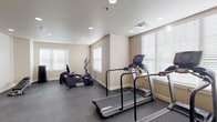 This fitness center is thoughtfully designed to support a well-rounded workout experience within a clean, functional layout. The space is organized into distinct zones, beginning with a dedicated cardio area housing multiple Landice treadmills equipped with digital display consoles, alongside elliptical cross-trainers and a stationary bike. Natural light filters in generously through several windows fitted with white horizontal blinds, creating a bright and inviting atmosphere throughout.

The flooring throughout the fitness center consists of durable dark tile, providing a practical and resilient surface suited to high-traffic exercise use. Recessed lighting is distributed evenly across the smooth white ceiling, supplementing natural light and ensuring consistent illumination at all hours.

A separate section of the space serves as a free weights and strength training area, featuring a full rack of dumbbells arranged neatly near the windows, along with an adjustable workout bench ideal for a variety of exercises. The neutral beige walls are accented with clean white baseboards, contributing to the space's modern yet understated aesthetic.

An additional open area provides generous room for floor exercises, stretching, or functional training. A door connects the two zones, allowing for smooth circulation through the facility. Overall, the fitness center presents a well-maintained, purpose-built environment designed to accommodate diverse fitness needs.