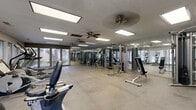 This fitness center offers a generously sized, single-floor workout facility designed to accommodate a comprehensive range of exercise activities. The space features durable large-format concrete flooring throughout, providing a sturdy and easy-to-maintain surface suited for heavy equipment use.

The strength training area is well-stocked with a variety of cable machines, weight stack units, a free-weight dumbbell rack, and adjustable benches, catering to users of all fitness levels. Full-length mirrors line the walls, visually expanding the space and allowing users to monitor their form during workouts.

The cardio zone is positioned along a wall of large windows, which flood the area with natural light and offer views of the outdoor surroundings, including what appears to be a pool area and landscaping. Two treadmills and multiple elliptical machines are positioned to take full advantage of these views. A set of French doors provides additional access and ventilation.

Overhead, recessed fluorescent panel lighting ensures the entire space is evenly and brightly illuminated. Ceiling fans are mounted throughout to provide consistent air circulation and comfort. The overall layout is open and functional, with ample floor space between equipment stations to allow for safe and unrestricted movement throughout the facility.