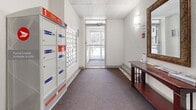 Located at 30 Campbell Court in Stratford, Ontario, this ground-floor common area serves as the primary entry and mailroom for the building. The space is efficiently laid out, combining functional and welcoming design elements in a compact footprint.

The entry side of the lobby features a durable speckled tile floor with a grid pattern, a sturdy metal security door equipped with a commercial-grade handle, and a ceiling-mounted security camera — all contributing to a secure and practical entryway.

Transitioning into the interior corridor, the flooring shifts to a muted, textured carpet that runs the length of the hall. A large bank of Canada Post parcel lockers and individual mailboxes lines one wall, providing organized mail and package reception for residents. A window at the end of the hallway allows natural light to filter in, offering a view of the surrounding neighbourhood and lending the corridor an open, airy feel.

On the opposite wall, a decorative ornate-framed mirror is mounted above a rich mahogany-toned console table, adding a touch of elegance to the utilitarian space. Wall-mounted sconce lighting provides warm, even illumination throughout. A baseboard heating unit runs along the lower wall, ensuring comfort during colder months. The textured white walls and drop-tile ceiling complete the clean, well-maintained aesthetic of this functional building lobby.