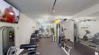 This fitness center is a fully outfitted exercise facility designed to support a comprehensive workout routine within a compact, efficiently organized footprint. The space features white walls accented by a bold yellow structural column, lending a clean and energetic aesthetic throughout. Track lighting runs along the ceiling, providing consistent, bright illumination across the entire room. Durable gray carpet flooring covers the workout area, offering traction and comfort underfoot.

The cardio section includes multiple treadmills and elliptical machines, arranged in a row and backed by large floor-to-ceiling mirrors that visually expand the space. Strength training is well represented, with a multi-station cable machine serving as a central fixture, complemented by a leg press machine and additional resistance equipment. A dedicated free weights area features a tiered dumbbell rack alongside a selection of medicine balls, catering to functional and strength-focused training alike.

A wall-mounted flat-screen television is positioned for easy viewing during workouts. Motivational "FITNESS" wall decals add to the gym's energetic atmosphere. A door at the rear provides access to the exterior, allowing for natural light and ventilation. The strategic use of mirrors throughout the space enhances depth and allows users to monitor their form, making this fitness center a practical and well-designed amenity.