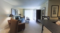 This single-floor residential unit offers a comfortable and functional layout designed to maximize livability. The living room serves as the central gathering space, featuring plush wall-to-wall carpeting that adds warmth underfoot. A large sliding glass door fitted with vertical blinds floods the room with natural light and provides access to the outdoors, creating an open and airy atmosphere. The living area comfortably accommodates a full seating arrangement, with ample space for a sofa, accent chair, and a central coffee table, making it well-suited for both relaxing and entertaining. Framed artwork adorns the walls, contributing to a welcoming ambiance. A textured ceiling adds a classic residential character throughout the space. Adjacent to the living room, a dining area with a dark-toned table and seating transitions seamlessly into the kitchen zone, supporting an open-concept flow between the spaces. Storage is addressed with a tall dark cabinet unit positioned near the sliding door. The neutral color palette of soft greens and warm beiges used throughout the unit creates a cohesive, calming aesthetic that makes the space feel inviting and move-in ready.