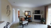 This single-floor residence offers a warm and inviting atmosphere, thoughtfully designed with a neutral color palette that creates a sense of calm throughout. The main living area serves as the heart of the space, featuring a built-in fireplace with decorative paneling that anchors the room and adds a classic architectural touch. A wall-mounted flat-screen television is positioned adjacent to the fireplace, making the area ideal for relaxed entertaining or quiet evenings at home. Soft, plush carpeting underfoot enhances the comfort of the living space, while the light beige walls contribute to an airy, open feel. A stylish console table, adorned with a table lamp and decorative ceramic accents in warm amber tones, adds character and a refined sense of style. A striped accent chair with an orange throw pillow provides a comfortable seating option and ties together the room's earthy color scheme. White-trimmed doorways lead to additional areas of the home, hinting at further well-appointed rooms beyond. The overall ambiance is one of understated elegance, blending functional design with tasteful décor to create a comfortable and cohesive living environment.