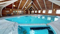 This single-level indoor recreational facility is defined by its striking architectural character, centered around a generously sized indoor swimming pool with crystal-clear blue water. The soaring vaulted ceiling, clad in warm natural wood planking and supported by bold white exposed beams, creates a chalet-inspired ambiance that sets the space apart. Globe pendant lights hang from the ceiling, casting a soft, inviting glow throughout.

The pool deck features distinctive large-format flagstone flooring that extends across the full perimeter, providing ample space for lounging with scattered bistro-style tables and chairs. A diving board is positioned along the pool's edge, and stainless steel entry rails offer convenient pool access.

The warm-toned cedar wood wall paneling is consistent throughout, complemented by a generous array of large windows that frame outdoor views and allow abundant natural light to fill the space. Along one wall, a glassed-in fitness room provides a dedicated workout area, while a cedar-paneled sauna room offers a private retreat. The facility also includes lounge and seating areas ideal for relaxing before or after a swim. The cohesive wood aesthetic, vaulted ceilings, and multifaceted amenities combine to create a welcoming and functional recreational environment.