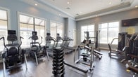 This fitness center offers a comprehensive workout environment designed to support a wide range of exercise routines. The space is finished with rich hardwood-style flooring and soft blue-gray walls accented by crisp white crown molding, creating a clean and motivating atmosphere. Recessed lighting throughout ensures consistent illumination across the entire floor.

The cardio zone is anchored by a row of treadmills and elliptical trainers, all equipped with individual digital monitors for tracking workout metrics. Large windows fitted with horizontal blinds line the walls, allowing ample natural light to pour in while providing views of the surrounding outdoors.

The strength training area is generously outfitted with a full rack of dumbbells in graduated weights, multiple cable and pulley machines, and a selection of Magnum-brand selectorized strength machines for targeted muscle training. An ab coaster and adjustable benches round out the functional training options available.

A full-length mirror wall expands the visual depth of the space and allows users to monitor their form during exercises. French doors provide a welcoming entryway, while an emergency exit ensures safety compliance. The overall layout is open and well-organized, offering ample space to move freely between equipment stations, making it suitable for both individual and simultaneous multi-user workouts.