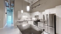 Located at 128 Mill St in Clayton, NC, this single-floor unit showcases a compelling blend of industrial and modern design across a well-appointed layout. Soaring ceilings with exposed ductwork and structural elements lend a loft-like character throughout, while smooth, polished concrete floors in a neutral grey-white tone unify the space with a clean, contemporary aesthetic.

The open-concept kitchen is a standout feature, outfitted with crisp white cabinetry, classic subway tile backsplash, dark granite countertops, and a full suite of stainless steel appliances including a gas range, dishwasher, and French door refrigerator. A substantial central island provides additional prep and gathering space. Pendant lighting suspended from above adds warmth and visual interest to the kitchen area.

The generously sized rooms feature tall walls in soft neutral tones and continue the polished floor aesthetic, creating bright, airy spaces ideal for a variety of uses. One room highlights an exposed white-painted brick accent wall, adding texture and historic character, along with access to natural light through tall windows.

The bathrooms are finished with white subway tile tub surrounds, dark countertop vanities, and clean modern fixtures — one bathroom is equipped with accessibility grab bars and a roll-in style tub. A dedicated laundry area houses a full-size front-loading washer and dryer. Built-in closet shelving provides practical storage throughout.