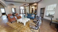 This single-floor space presents an open, well-appointed interior designed for comfort and community. The main lounge area commands attention with its soaring ceilings and a striking circular chandelier that anchors the room. The seating arrangement features a mix of a blue upholstered sofa accented with coral pillows, rust-toned armchairs, and sleek metal-framed chairs, all centered around a decoratively carved white coffee table atop a warm gold-toned area rug. Tile flooring runs throughout, adding durability and a clean aesthetic.

An elegant arched doorway connects the lounge to an adjacent dining or gathering area, which benefits from abundant natural light streaming through large windows and glass doors. A textured accent wall frames the transition to a secondary space that appears to include a kitchen or kitchenette, illuminated by pendant lighting and equipped with visible appliances. Additional recessed hallways suggest further functional areas nearby. The overall ambiance is warm and inviting, blending contemporary and transitional design elements to create a polished, cohesive communal environment.