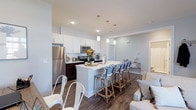 Located at 6770 Printers Blvd in New Albany, Ohio, this single-floor residence offers a thoughtfully designed open-concept layout that seamlessly connects the kitchen, dining, and living areas. The kitchen is a standout feature, showcasing crisp white shaker cabinetry, rich dark wood lower cabinets, and stainless steel appliances, including a full-size refrigerator, range, and microwave. A large central island with a light-toned countertop serves as the heart of the space, providing ample prep area and casual seating with stylish blue-and-white bistro-style barstools. Pendant lighting hangs above the island, adding both function and visual interest. The flooring throughout is a warm, wood-tone hardwood, lending continuity and a grounded feel to the open floor plan. Neutral light gray walls complement the clean, contemporary aesthetic and create a bright, airy atmosphere. Recessed lighting and crown molding details add a polished finishing touch to the ceilings. The living area flows naturally from the kitchen, creating an inviting and cohesive space ideal for both everyday living and entertaining. The overall design balances modern style with comfortable, functional living.