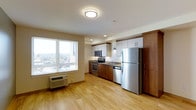 This well-appointed single-floor space offers a clean, modern design that maximizes both functionality and comfort. The open-concept layout seamlessly connects the kitchen and living area, creating a bright and welcoming atmosphere.

The kitchen is a standout feature, equipped with a full suite of stainless steel appliances including a refrigerator, electric range, built-in microwave, and dishwasher. Cabinetry combines warm wood-toned lower cabinets with crisp white upper cabinets, offering generous storage throughout. A dark stone-look countertop adds contrast and a touch of sophistication to the workspace.

Light-toned hardwood flooring runs throughout the space, lending warmth and a sense of continuity. The living area benefits from a large picture window that floods the interior with natural light and frames an elevated outdoor view, suggesting an upper-floor position with scenic surroundings.

Recessed and flush-mount ceiling lighting provides ample illumination, while a wall-mounted HVAC unit ensures year-round climate control. The overall ambiance is clean, contemporary, and move-in ready, making it a practical and attractive space for everyday living.