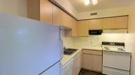 This single-floor space is thoughtfully laid out to maximize functionality and comfort. The kitchen stands out as a well-organized area featuring light wood cabinetry that extends to the upper walls, providing generous storage space. The countertops offer ample room for meal preparation, complemented by a stainless steel sink and a suite of white appliances including a refrigerator, dishwasher, and range. A built-in microwave is conveniently mounted above the counter, adding to the kitchen's practicality. The cabinetry's warm, natural tones create a clean and welcoming atmosphere, while the dark hardwood flooring adds a grounding contrast to the lighter elements throughout. Overhead lighting illuminates the space effectively, and a ceiling vent ensures comfortable air circulation year-round. The L-shaped kitchen layout is designed to optimize workflow, making it an efficient and user-friendly space for cooking and entertaining. Overall, the space presents a straightforward, functional design with attention to storage and utility, making it well-suited for everyday living.