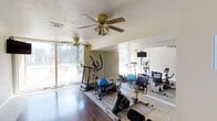 This dedicated fitness space is thoughtfully designed to support a complete workout experience. The room features hardwood flooring complemented by a neutral area rug, providing a comfortable and durable surface for exercise equipment. A variety of cardio machines — including an elliptical trainer, stationary bike, and treadmill — are arranged throughout the space, along with a stability ball and additional seating for stretching or rest. Full-length wall mirrors run along one side of the room, amplifying the sense of space while allowing users to monitor their form during workouts. A mounted screen is positioned above the mirror for on-demand fitness content or entertainment. The room is well-ventilated with two ceiling fans, ensuring comfort during intense exercise sessions. Large sliding glass doors along the opposite wall flood the space with natural light and offer a pleasant view of an outdoor pool and patio area, creating an open, airy atmosphere. The ceiling has a textured finish, and the overall color palette of warm beiges and neutrals contributes to a clean, functional environment. This single-floor fitness space blends practicality with comfort, making it an ideal setting for daily exercise routines.