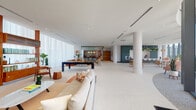 Located along Biscayne Boulevard in Miami's vibrant Upper East Side, this single-floor amenity space delivers a refined, open-plan environment designed for both relaxation and social engagement. The expansive layout is anchored by polished terrazzo flooring throughout, which reflects natural light flooding in from floor-to-ceiling glass walls and sheer curtains that frame sweeping views of the Miami skyline and lush greenery below.

The main lounge area features multiple distinct seating arrangements, including a tan leather sofa paired with natural rattan chairs and warm wood accent tables, creating an inviting, mid-century-modern aesthetic. Adjacent to the lounge, a dedicated game room houses a full-sized billiards table alongside shelved bookcases and additional casual seating. A curved dining and café area benefits from dramatic curved floor-to-ceiling windows, filling the space with abundant natural light and offering panoramic treetop views.

The sleek, contemporary kitchen is outfitted with matte teal cabinetry, integrated appliances, and a clean white countertop with a brass-finished sink. A co-working zone features a comfortable sectional sofa, a wall-mounted television, and glass-partitioned rooms for focused work or private meetings. Smooth white cylindrical columns punctuate the space, while lush indoor plants add a fresh, organic element throughout.