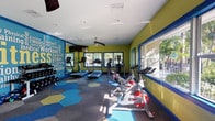 This single-floor fitness center is designed with energy and functionality in mind, offering a comprehensive workout environment for all fitness levels. The space is immediately striking thanks to its bold color palette — chartreuse-yellow walls accented by vivid cobalt blue trim and window frames — creating an atmosphere that feels motivating and dynamic.

The cardio zone is well-appointed, featuring multiple treadmills and elliptical cross-trainers lined along one wall, complemented by a set of spin bikes positioned near large, blue-framed windows. These oversized windows flood the space with abundant natural light and offer views of lush palm trees and greenery outside, enhancing the workout experience with a connection to the outdoors.

On the opposite end of the room, a multi-station cable strength machine anchors the strength-training area, accompanied by free weights and a weight bench. Full-length mirrors line this section of the wall, visually expanding the space while allowing users to monitor their form.

Two ceiling fans circulate air throughout the room, and a mounted flat-screen television adds entertainment value. The floor features a distinctive patterned carpet with bold hexagonal shapes in blue, teal, and gold, adding a playful, modern design element. Motivational fitness-themed word art murals decorate the walls, reinforcing the space's energetic and health-focused identity.