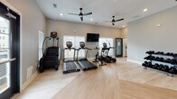 Located at 10 Shell Creek Dr in Port Royal, South Carolina, this dedicated fitness center is designed to support a comprehensive workout experience within a single, well-organized floor. The space features light hardwood flooring laid in a herringbone pattern, which lends a clean, modern aesthetic throughout.

The cardio zone is generously appointed with multiple treadmills, an elliptical trainer, a stair climber, and a stationary bike — all from True Fitness — arranged to maximize open floor space. A wall-mounted flat-screen television is positioned to keep exercisers entertained, while multiple ceiling fans with sleek, dark finishes ensure consistent airflow and comfort during workouts.

The strength training area is equally well-stocked, featuring a multi-functional cable and resistance machine, a dedicated adjustable bench, and a tiered dumbbell rack with a full range of free weights. Full-length mirrors line one wall, creating a sense of expanded space and allowing users to monitor their form during exercises.

Natural light fills the room through multiple windows fitted with white horizontal blinds, complementing the recessed lighting overhead. The neutral gray and warm beige walls contribute to a calm, focused atmosphere. Access is provided through both interior and keyed exterior doors, offering convenient entry from outdoors.