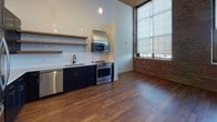 Located at 22 Merrimack in Lawrence, MA, this single-floor unit showcases a compelling blend of industrial heritage and contemporary design. Soaring ceilings with exposed ductwork and wood-paneled soffits lend the space a loft-like character, while rich hardwood flooring flows consistently throughout, grounding the open layout.

The kitchen is a standout feature, equipped with navy blue cabinetry, stainless steel appliances including a gas range, built-in microwave, refrigerator, and dishwasher, complemented by open wood shelving and a classic white subway tile backsplash. A stainless steel hood vent adds an industrial touch.

The living area benefits from oversized windows fitted with roller shades, allowing generous natural light while framing views of the surrounding neighborhood. Original exposed brick walls serve as a defining architectural element throughout the space.

A dedicated in-unit stacked washer and dryer offers added convenience. The walk-in closet provides ample storage with adjustable shelving and hanging rods. The bathroom is finished with hexagonal gray floor tiles, a wood-toned vanity, brass fixtures, white subway tile walls, and a glass-enclosed walk-in shower, creating a clean, modern aesthetic.
