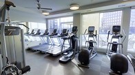 This fitness center is designed to provide a comfortable and functional workout environment. The space is lined with floor-to-ceiling windows along two walls, flooding the room with natural light and offering views of the surrounding urban landscape. A row of treadmills is positioned facing the windows, allowing users to enjoy the outdoor scenery during their workouts. Elliptical trainers and an exercise bike are also available, offering a variety of cardio options. The flooring consists of durable, cushioned gym-grade material in a neutral tone, providing both comfort and practicality. Ceiling fans are mounted overhead, helping to maintain air circulation throughout the space. Recessed and flush-mount lighting supplements the natural light, ensuring the room remains well-lit at all times. Mirror panels are incorporated into the design, visually expanding the space and allowing users to monitor their form. The overall ambiance is clean, modern, and well-maintained, with equipment arranged to maximize usability and movement within the room. The combination of large windows, quality equipment, and thoughtful layout makes this fitness center a well-rounded facility for cardiovascular training.