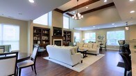 This beautifully appointed single-floor space exudes a refined, welcoming ambiance through its thoughtful architectural design and high-end finishes. The centerpiece of the main living area is a classic fireplace flanked by rich, dark wood built-in bookshelves that extend toward the ceiling, offering both display and storage space. The soaring two-story ceilings, accented by elegant dark wood coffered beams, lend an impressive sense of grandeur and openness to the room. An elegant chandelier hangs overhead, complementing the recessed lighting that ensures the space is warmly and evenly lit. Large windows along the walls invite generous amounts of natural light, brightening the neutral, warm-toned walls and dark hardwood flooring throughout. The living area is furnished with plush, cream-colored sofas and armchairs arranged around a central coffee table atop a soft area rug, creating an inviting setting for relaxation and conversation. Adjacent to the main seating area, a dining space with chairs and a table provides a natural transition for gathering and entertaining. The overall aesthetic blends traditional craftsmanship with comfortable, modern furnishings, resulting in a space that feels both sophisticated and effortlessly livable.