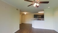 This single-story space features an open-concept layout that seamlessly connects the living area to the kitchen. Rich, dark hardwood-style flooring runs throughout, lending warmth and a polished aesthetic to the interior. Crown molding accents the ceiling, adding a refined architectural detail, while a ceiling fan with integrated lighting anchors the living area. The kitchen is well-appointed with dark espresso cabinetry, granite-look countertops, and stainless steel appliances including a microwave and range. A peninsula-style counter extends into the living space, creating a natural divide between the two areas while maintaining the open flow. The kitchen benefits from its own overhead lighting, ensuring the workspace is well-illuminated. Neutral, light-toned walls complement the darker flooring and cabinetry throughout. A hallway visible toward the rear of the living area leads to additional rooms, suggesting a functional layout beyond the main living space. Overall, the space presents a clean, contemporary feel with a practical and cohesive design.