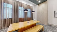 This single-floor professional space is designed to support both collaborative work and private meetings, combining contemporary aesthetics with functional amenities. The main lounge area is anchored by a long natural wood communal table with matching benches, illuminated by a pair of striking navy blue pendant lights that hang from a tray ceiling with a deep teal accent. Floor-to-ceiling draping curtains frame large windows, allowing soft natural light to filter through sheer underlays, creating a warm yet refined atmosphere.

The flooring throughout features a distinctive grey and white patterned carpet that adds visual texture and continuity across the space. Light wood wall paneling and white tile-mosaic accent walls contribute to a clean, upscale interior palette.

A private conference room, enclosed by black-framed glass doors, offers a more intimate setting with a rectangular wood table, ergonomic rolling chairs, a wall-mounted display screen, and a sleek pendant light overhead. Decorative wall art adds character to the room.

Adjacent transitional areas include a welcoming alcove with a white upholstered chair and side table set against a textured mosaic wall, as well as an open corridor showcasing framed artwork and built-in access panels. The overall ambiance is polished and professional, well-suited for meetings, co-working, or community gatherings.
