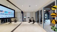 This single-level fitness facility is thoughtfully designed to accommodate a focused workout experience within a sleek, modern aesthetic. The cycling area serves as the centerpiece of the space, featuring multiple stationary bikes arranged on a light-toned, textured floor that adds both style and durability underfoot. A large floor-to-ceiling mirror spans one wall, amplifying the sense of openness while allowing users to monitor their form during exercise. Recessed lighting in the white ceiling provides bright, even illumination throughout the room, contributing to a clean and energizing atmosphere. A wall-mounted display screen is integrated near the cycling stations, offering the ability to stream classes or track workout metrics. An in-wall climate control panel ensures a comfortable environment during intense sessions. Framed by sleek aluminum-framed glass partitions, an adjoining equipment room is visible from the cycling area, housing additional strength and cardio equipment to support a comprehensive fitness routine. The overall design emphasizes functionality and cleanliness, with a neutral white and navy color palette that creates a professional and motivating environment.