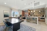 Located at 2235 Treehouse Ln in Corona, California, this thoughtfully designed single-floor community space combines recreation, dining, and lounge functionality in one cohesive open-plan layout. The interior is finished with large-format light tile flooring throughout, creating a clean and bright foundation that complements the warm, contemporary aesthetic.

The kitchenette area is a standout feature, showcasing wood-tone cabinetry, a white quartz countertop island with seating for two, open wood-clad shelving, a beverage refrigerator, a microwave, and a sink. A bold wood-plank accent wall provides a rich backdrop, while a trio of globe pendant lights suspended from brass ring fixtures add a designer touch overhead.

Adjacent to the kitchenette, the dining zone features a dark wood table paired with vivid teal chairs, anchored by a patterned area rug and illuminated by an artistic gold wire pendant light. A wall-mounted flat-screen television and large windows with sheer curtains allow natural light to fill the space.

The recreational corner includes a full shuffleboard table flanked by colorful landscape artwork, adding a casual, social atmosphere. A bold geometric-patterned accent wall near the lounge seating area injects personality and visual contrast. The overall ambiance is modern and inviting, blending eclectic art with warm natural materials.