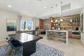 Located at 2235 Treehouse Ln in Corona, California, this thoughtfully designed single-floor community space combines recreation, dining, and lounge functionality in one cohesive open-plan layout. The interior is finished with large-format light tile flooring throughout, creating a clean and bright foundation that complements the warm, contemporary aesthetic.

The kitchenette area is a standout feature, showcasing wood-tone cabinetry, a white quartz countertop island with seating for two, open wood-clad shelving, a beverage refrigerator, a microwave, and a sink. A bold wood-plank accent wall provides a rich backdrop, while a trio of globe pendant lights suspended from brass ring fixtures add a designer touch overhead.

Adjacent to the kitchenette, the dining zone features a dark wood table paired with vivid teal chairs, anchored by a patterned area rug and illuminated by an artistic gold wire pendant light. A wall-mounted flat-screen television and large windows with sheer curtains allow natural light to fill the space.

The recreational corner includes a full shuffleboard table flanked by colorful landscape artwork, adding a casual, social atmosphere. A bold geometric-patterned accent wall near the lounge seating area injects personality and visual contrast. The overall ambiance is modern and inviting, blending eclectic art with warm natural materials.