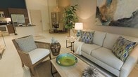 This thoughtfully designed single-floor residence offers an open and airy layout that encourages effortless flow between living and dining areas. The living room serves as the heart of the home, anchored by a plush neutral-toned sofa accented with vibrant blue and green patterned throw pillows, a light wood coffee table, and a cozy armchair that invite relaxed conversation. A bold statement bowl in emerald green adds a pop of color atop the coffee table, complemented by eclectic décor accents including a sculptural centerpiece and ceramic accessories in muted coastal tones. A tall, lush fiddle-leaf fig tree brings a refreshing natural element to the interior, while warm ambient lighting from a classic table lamp creates an inviting atmosphere. Abstract wall art in earthy and oceanic hues adds artistic character to the space. Just beyond the living area, a dining space is visible, furnished with a round table and chairs, offering an intimate setting for meals. Light-toned flooring runs throughout, enhancing the sense of openness and continuity across the floor plan. The overall ambiance is warm, curated, and contemporary, reflecting a refined yet approachable aesthetic.