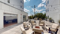 This outdoor amenity space is set within the interior courtyard and extended terrace of a contemporary multi-story building clad in clean white paneling. The layout is divided into distinct functional zones, each designed to encourage gathering and relaxation. At the heart of the courtyard sits a central fire pit surrounded by cushioned lounge chairs, creating an inviting focal point for socializing. String lights are strung overhead, providing ambient illumination that enhances the space during evening hours.

Picnic-style dining tables and café bistro sets are distributed throughout the patio, offering flexible seating arrangements for both casual and more structured gatherings. Large planters with greenery add natural elements and visual interest against the building's modern facade. A garage-style door opens from an interior amenity room directly onto the courtyard, seamlessly connecting indoor and outdoor spaces.

The extended terrace portion features additional seating with open views of the surrounding urban landscape, framed by black metal railings and mature trees beyond the perimeter. Large-format tile paving runs throughout the entire space, providing a cohesive and polished finish. The open-sky setting ensures ample natural light throughout the day, contributing to an inviting and well-appointed outdoor environment.