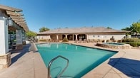 Located at 9270 Eagle Ranch Rd NW in Albuquerque, New Mexico, this single-level outdoor amenity area is designed to take full advantage of the sunny high-desert climate. The centerpiece of the space is a generously sized swimming pool with clear turquoise water, surrounded by a broad concrete deck furnished with multiple lounge chairs and shaded umbrellas — ideal for relaxing and sunbathing. A raised spa with stone surround sits adjacent to the main pool, offering a dedicated area for hydrotherapy and leisure. A handsome wood-beamed pergola provides a shaded outdoor living area outfitted with comfortable seating, perfect for socializing or unwinding out of the sun. A community building with stone-accented stucco exterior lines one side of the pool deck, housing additional amenities and facilities. The grounds are thoughtfully landscaped with mature shade trees, manicured hedges, and water-conscious xeriscape featuring decorative gravel and native plantings. Gated entry with iron fencing ensures a secure and private environment. The overall ambiance reflects the classic Southwestern architectural aesthetic, blending natural stone, earth tones, and lush greenery against the backdrop of Albuquerque's characteristic bright blue skies.