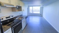 This single-floor space presents a clean, modern aesthetic defined by an open-plan layout that seamlessly connects the kitchen and living area. The kitchen is equipped with a stainless steel sink, black countertops, a gas range, and a dishwasher, complemented by light wood upper cabinetry and an integrated range hood. Smooth, neutral-toned flooring extends throughout the entire space, lending a cohesive and contemporary feel underfoot. The living area is bright and airy, anchored by a large picture window that floods the interior with natural light and frames views of the surrounding urban streetscape. The white walls and minimal design language keep the space feeling open and versatile. A floor-mounted heating vent beneath the window ensures year-round comfort. The overall ambiance is functional and modern, ideal for those who appreciate clean lines, an uncluttered environment, and a well-connected open-plan living experience. The layout makes efficient use of the available space while maintaining a sense of openness and flow between the kitchen and living zones.