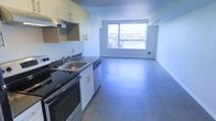 This single-floor space presents a clean, modern aesthetic defined by an open-plan layout that seamlessly connects the kitchen and living area. The kitchen is equipped with a stainless steel sink, black countertops, a gas range, and a dishwasher, complemented by light wood upper cabinetry and an integrated range hood. Smooth, neutral-toned flooring extends throughout the entire space, lending a cohesive and contemporary feel underfoot. The living area is bright and airy, anchored by a large picture window that floods the interior with natural light and frames views of the surrounding urban streetscape. The white walls and minimal design language keep the space feeling open and versatile. A floor-mounted heating vent beneath the window ensures year-round comfort. The overall ambiance is functional and modern, ideal for those who appreciate clean lines, an uncluttered environment, and a well-connected open-plan living experience. The layout makes efficient use of the available space while maintaining a sense of openness and flow between the kitchen and living zones.