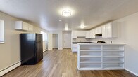 This studio unit at 645 Wolff St in Denver, Colorado offers a practical and well-organized single-floor layout. The open-concept kitchen and living area form the heart of the space, finished with warm wood-look laminate flooring that runs throughout. The kitchen is outfitted with crisp white cabinetry — both upper and lower — paired with dark countertops, a gas range, a full-size refrigerator, and an under-cabinet range hood. A built-in peninsula with open shelving below provides a natural divide between the kitchen and the living area, adding both storage and visual interest. The living area is bright and open, with multiple windows fitted with blinds that allow natural light to fill the room. A baseboard heating system runs along the walls, providing consistent warmth. The textured ceiling adds a classic residential character throughout the space. The bathroom features a corner vanity with a white cabinet base, dark countertop, single basin sink, and a large wall mirror illuminated by overhead lighting, set on tile flooring. A dedicated closet offers convenient built-in storage. The neutral color palette and clean finishes create a functional and cohesive atmosphere throughout this compact Denver studio.