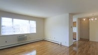 This single-floor space offers a clean, functional layout designed to maximize comfort and usability. The living area is a standout feature, showcasing polished hardwood floors that reflect natural light streaming in through a large window, creating a bright and airy atmosphere. Neutral white walls throughout provide a versatile backdrop, making the space feel open and easy to personalize. A baseboard heating system runs along the walls, ensuring efficient climate control, while a wall-mounted air conditioning unit adds year-round comfort. The living area flows naturally into the kitchen, which is visible through an open passage and features warm-toned wood cabinetry that adds a touch of warmth to the otherwise light and neutral palette. The kitchen includes ample cabinet storage and appears well-organized for everyday use. The overall ambiance of the space is modest yet practical, with clean finishes and a straightforward design that prioritizes function. The gleaming hardwood floors tie the living and kitchen areas together cohesively, lending a timeless quality to the interior. This space is well-suited for those seeking a comfortable, low-maintenance environment with classic details.